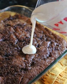 a person pours milk on top of a brown cake in a glass dish with yellow cloth