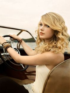 a beautiful blonde woman sitting in the driver seat of an old fashioned car with her hand on the steering wheel