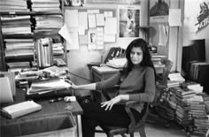 a woman sitting at a desk in an office with stacks of papers on the walls