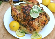 a whole chicken with lemons and rosemary garnish on a white plate next to a sliced lemon