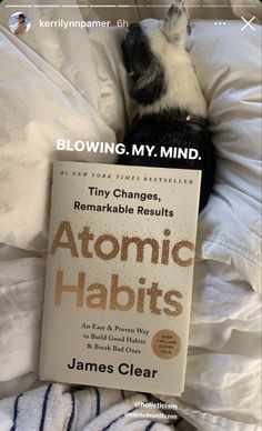 a dog laying on top of a bed next to a book titled atomic habitts