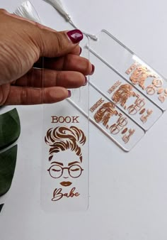 a person is holding up some bookmarks with their faces on them and the words book are in gold foil