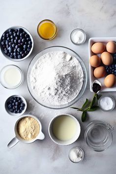 ingredients for blueberry cake laid out in bowls on a white counter top, including eggs, milk, and flour