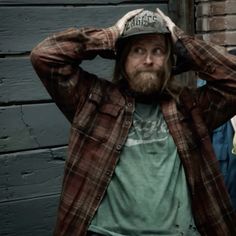 a man with a beard wearing a plaid jacket and hat standing in front of a brick wall