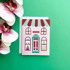 there is a card with a small store on it and flowers in front of it