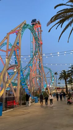 people are walking around an amusement park at night with lights strung above the roller coaster