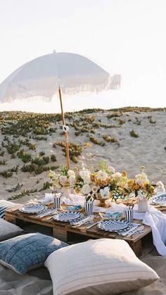 a table set up on the beach with an umbrella over it and flowers in vases