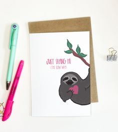 a card with a sloth hanging on a tree branch, next to a pen and paper clip