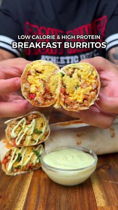 a person holding two burritos on top of a wooden table next to a bowl of ranch dressing