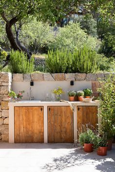 an outdoor kitchen with wooden cabinets and potted plants on the outside wall, surrounded by greenery