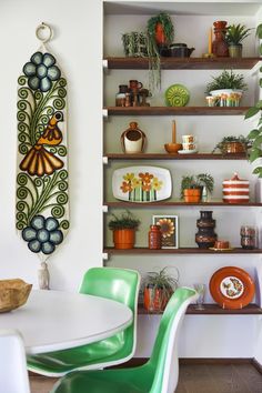 a dining room table with green chairs and plates on the wall behind it, surrounded by shelves filled with potted plants