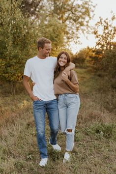 a man and woman are walking through the grass together in front of some trees with their arms around each other