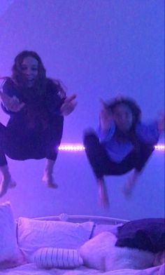 two women are jumping in the air on a bed with pillows and lights behind them