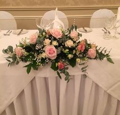 the table is set with white linens, pink roses and greenery on it