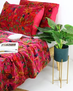 a bed with red sheets and pillows next to a plant in a pot on a gold stand