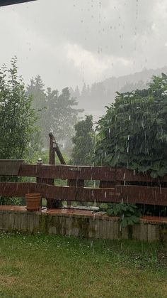 the rain is coming down on some benches in the yard with trees and bushes behind them