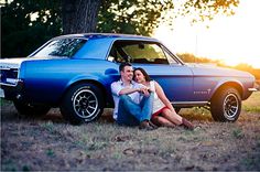 a man and woman sitting in front of a blue car with the sun setting behind them