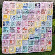 a colorful quilt hanging on the side of a brick wall in front of some bushes