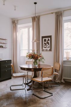 a dining room table with chairs and a vase on top of it in front of two windows