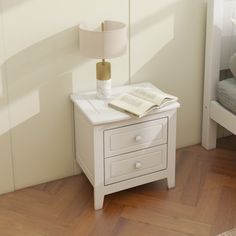 a white nightstand with a book on it next to a lamp and bed in a room