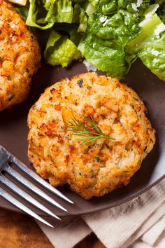 two crab cakes on a plate next to a salad with a fork and knife in the foreground