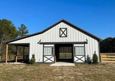 a white barn with two doors on the front and side of it, surrounded by trees