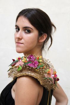 a woman with flowers on her neck wearing a black dress and gold necklace, standing in front of a white wall