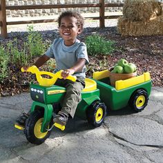 a young boy is riding a green and yellow toy car with apples in the back