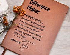 a brown book with writing on it sitting next to some flowers and an orange flower