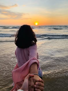 two people hold hands as the sun sets over the ocean on a beach in front of them