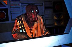 an animated character in a sci - fi setting with lights on, and other electronic equipment behind him