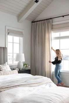 a woman standing on the edge of a bed in front of a window with curtains