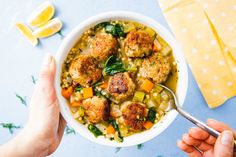 two hands holding a bowl of soup with meatballs and vegetables on the table next to lemons