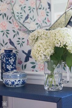 white flowers in a vase on a blue table next to a mirror and wallpaper