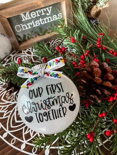 a christmas ornament with the words our first christmas together on it next to pine cones and red berries