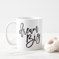 a white coffee mug with the words dream big on it next to a donut