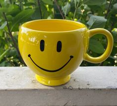 a yellow mug with a smiley face painted on the side sitting on a ledge in front of some bushes