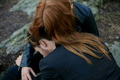 a woman with red hair sitting on the ground next to a man in black jacket