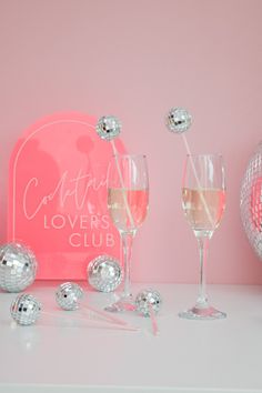 two wine glasses and some disco balls on a table next to a pink sign that says confection lover's club