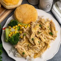 a white plate topped with chicken, broccoli and rice next to bread rolls