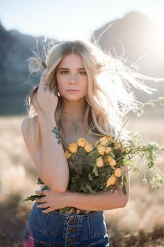 a woman with long blonde hair and blue jeans is holding yellow roses in front of her face