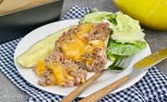 a white plate topped with meat covered in cheese next to a knife and fork on top of a checkered table cloth