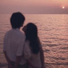 a man and woman standing on the beach looking out at the ocean as the sun sets