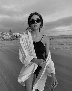 a woman standing on top of a beach next to the ocean wearing sunglasses and a black bathing suit