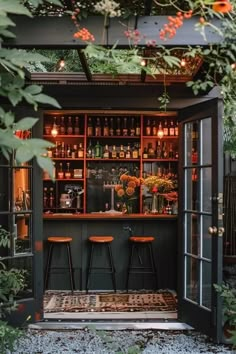 an outdoor bar with two stools and bottles on the back wall, surrounded by greenery