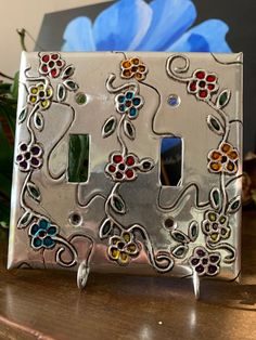 a decorative light switch cover with flowers and vines painted on the side, sitting on a table next to a blue flower