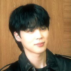 a young man with black hair wearing a leather jacket and looking at the camera while standing in front of a wooden wall