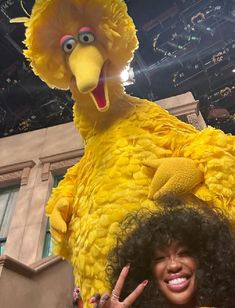 a woman standing next to a large bird mascot at a convention or show with her hand up in front of her face