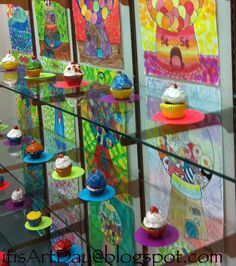 colorful cupcakes are on display in front of glass cases with paintings behind them