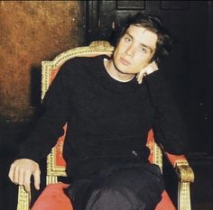 a man sitting on top of a red chair next to a wooden door and wearing a black sweater